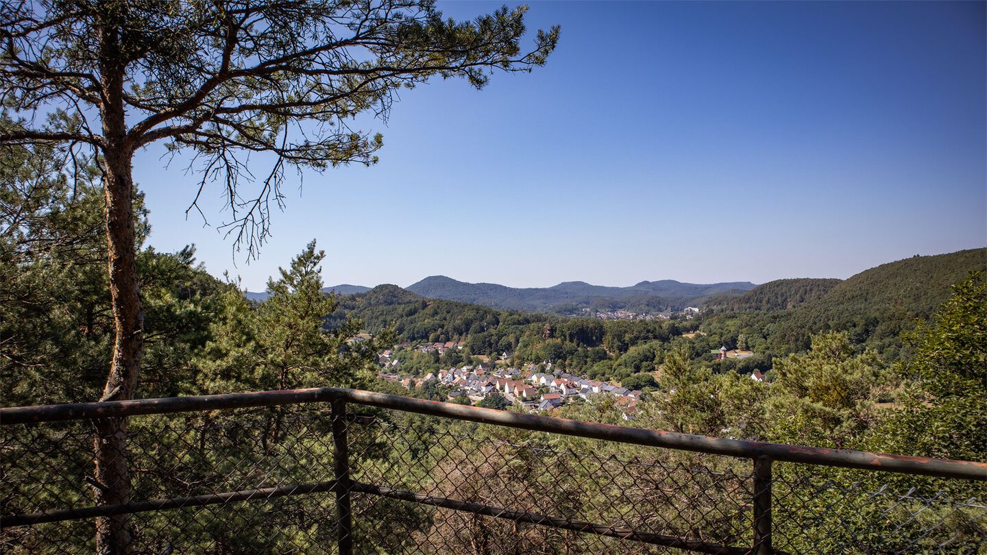Rastplatz Dorfblick am Hahnberg mit Geländer und Kiefer im Vordergrund, Blick über Erfweiler und den Wasgau bei klarem Himmel