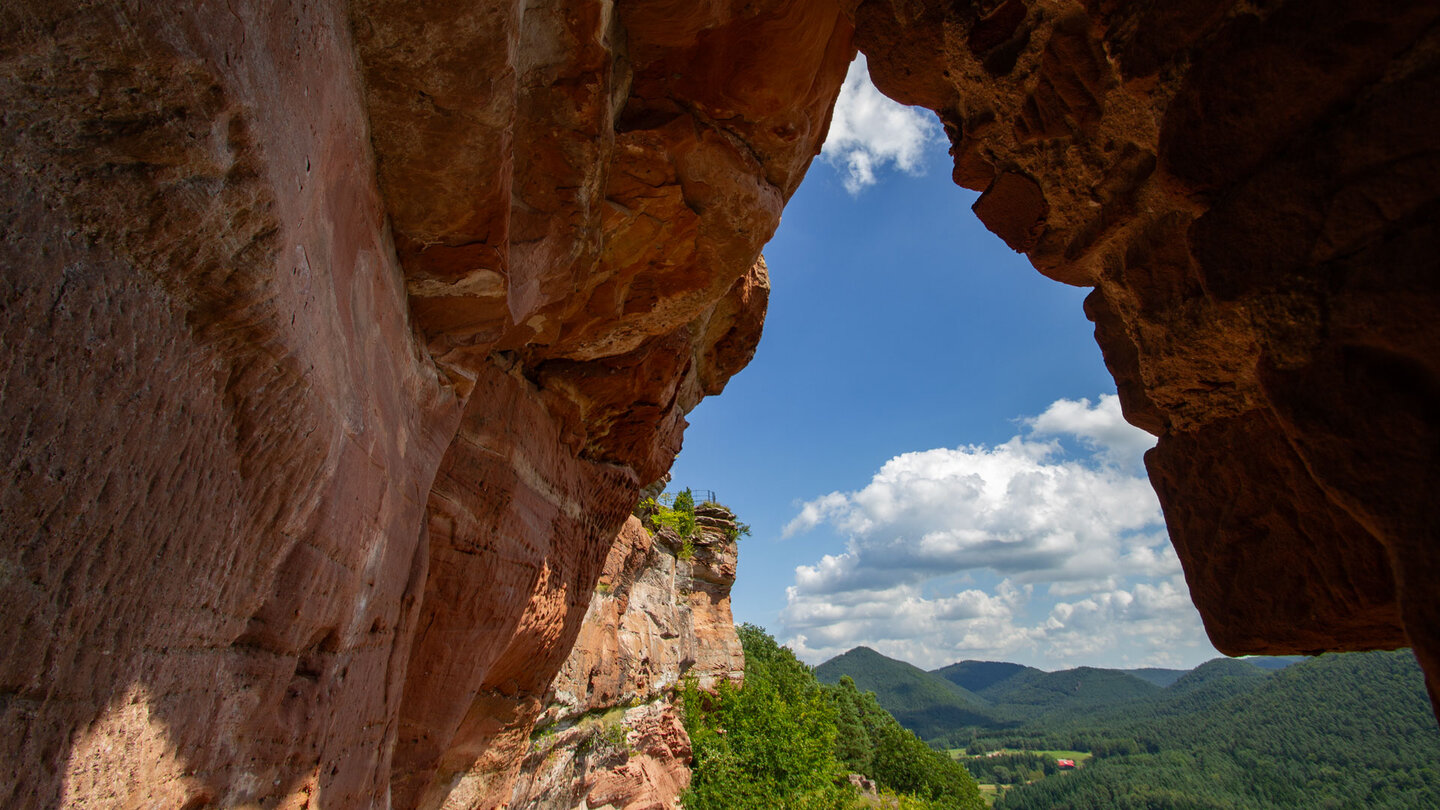 Felsdurchblick auf der Dahner Burgengruppe, rötliche Buntsandsteinwände rahmen den Blick auf einen Felspfeiler und die Höhenzüge des Wasgaus