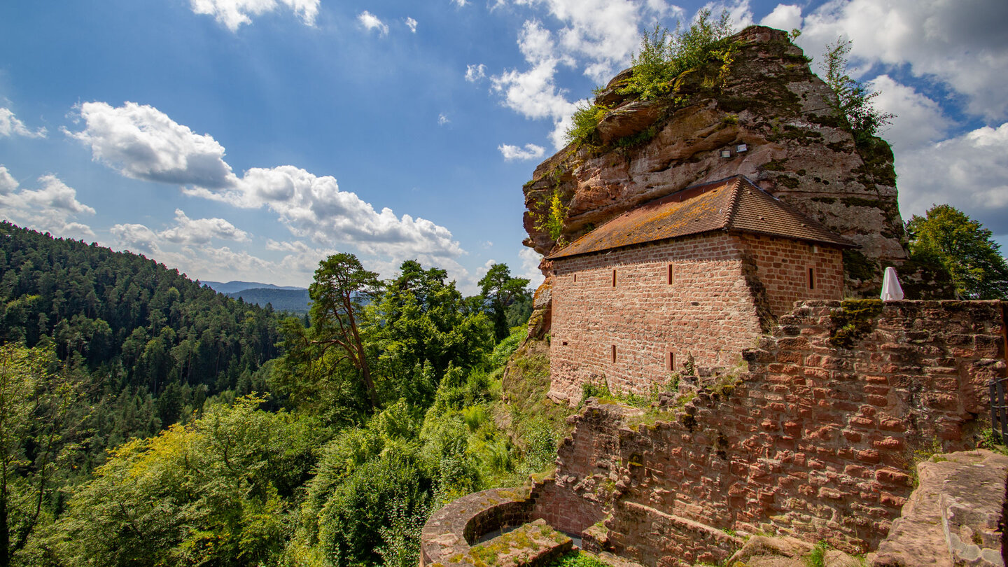 Rekonstruiertes Stallgebäude mit Ziegeldach am Buntsandsteinfelsen der Burgruine Grafendahn, bewaldete Berge im Hintergrund