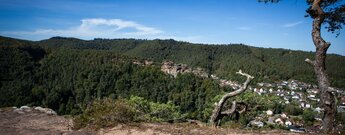 Blick vom Felsplateau des Kahlen Felsens über Hauenstein im Tal auf den Burghalder Felsen als rötliche Felswand im bewaldeten Hang unter blauem Himmel
