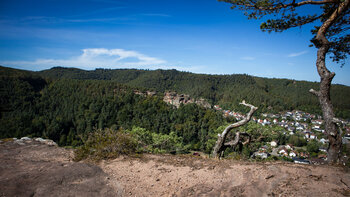 Blick vom Felsplateau des Kahlen Felsens über Hauenstein im Tal auf den Burghalder Felsen als rötliche Felswand im bewaldeten Hang unter blauem Himmel