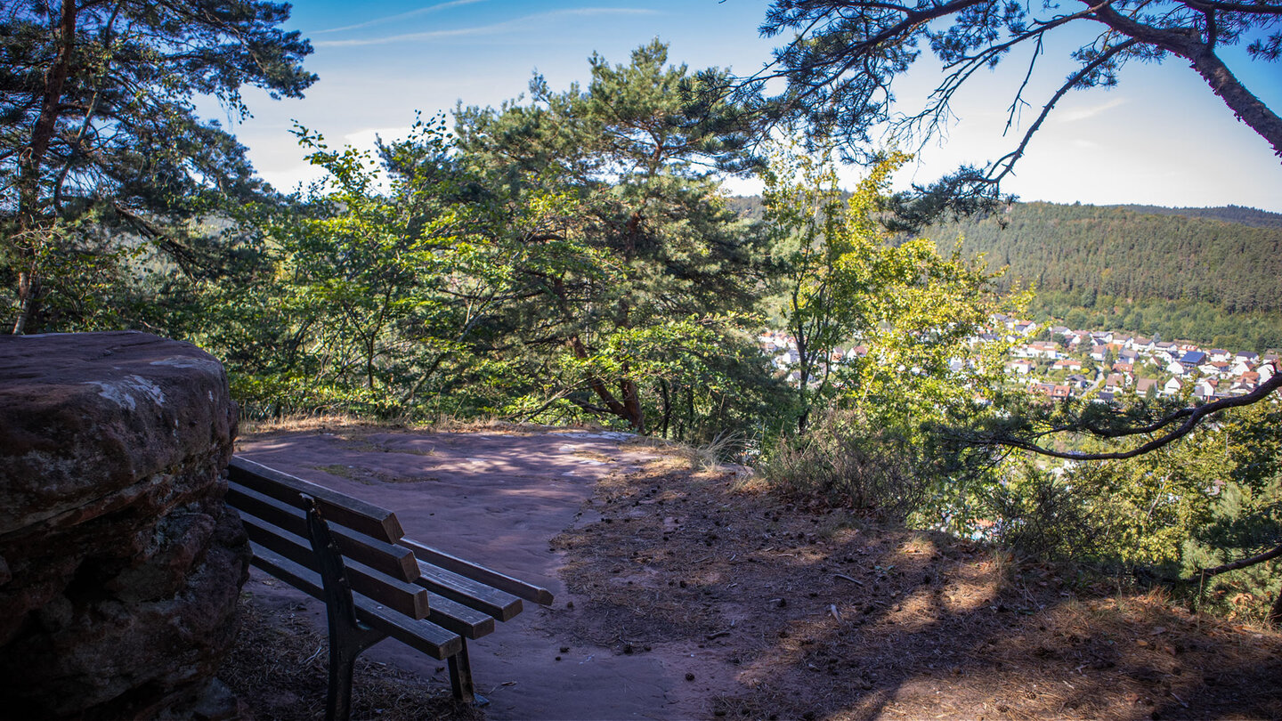 Holzbank an einem Buntsandsteinfels mit Blick durch Kiefern und Eichen auf Hauenstein mit roten Dächern und bewaldete Bergrücken