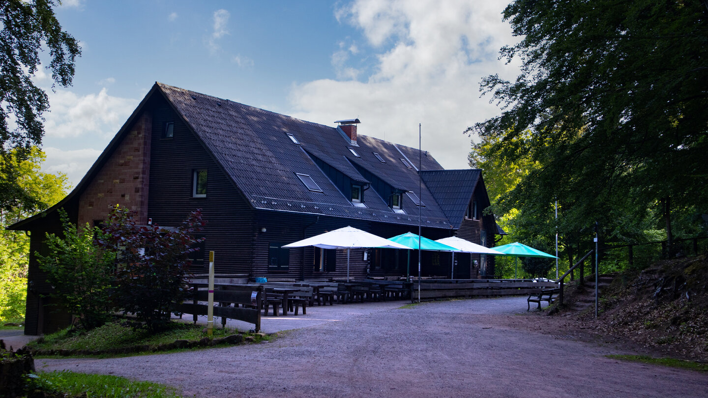 Wanderheim Dicke Eiche – großes Holzhaus mit dunkler Fassade, Terrasse und Sonnenschirmen auf einer Lichtung im Pfälzerwald
