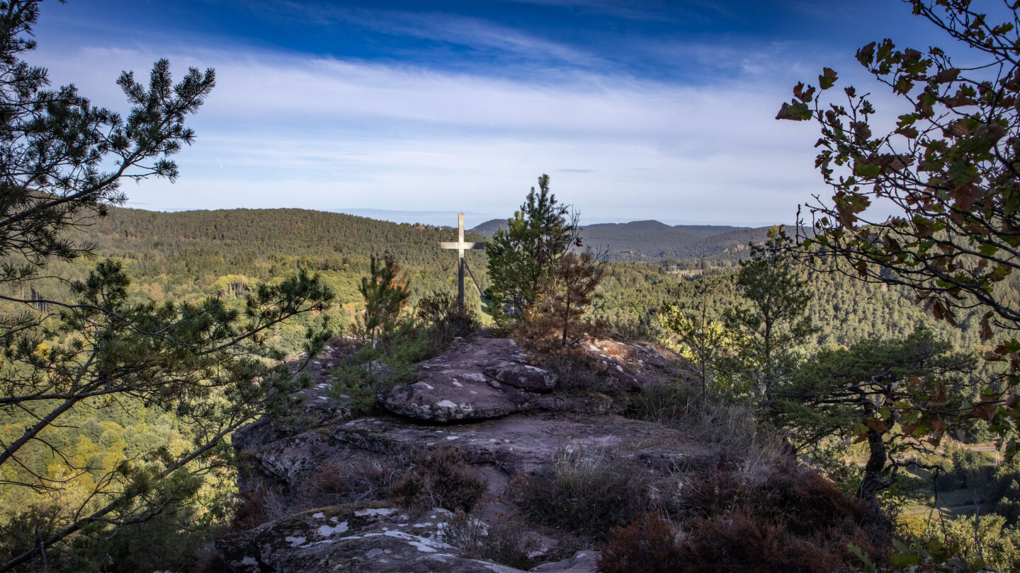 Felsiges Plateau des Hockersteins mit Gipfelkreuz und jungen Kiefern, dahinter weite bewaldete Bergrücken unter blauem Himmel mit Schleierwolken