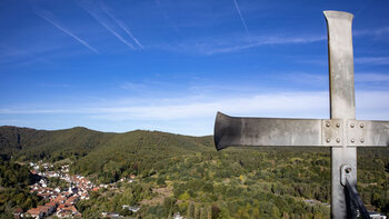 Metallenes Gipfelkreuz am Hockerstein in Nahaufnahme, dahinter Blick auf Schwanheim mit Kirchturm und bewaldete Hügel unter blauem Himmel