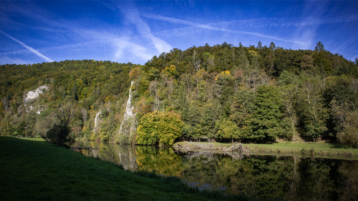 Bewaldeter Felsen mit herbstlich gefärbten Bäumen spiegelt sich im ruhigen Wasser der Donau